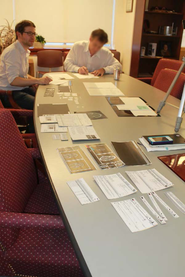 Russell Cornwell moderates a complicated paper prototype evaluation with an influential radiologist while both parties sit in a hospital office at the end of a long conference table strewn with paper prototype components